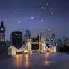 A miniature model of the Tower Bridge in London is illuminated at night, with a blurred cityscape and Ferris wheel in the background. Anteprima dell’immagine prodotto