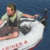 A man is sitting in an inflatable boat on the water, holding onto an outboard motor. Produktbild-Vorschau