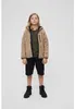 A young person stands facing forward against a plain white background, wearing a beige fleece jacket over a green t-shirt and black shorts, with black boots. Produktbild-Vorschau