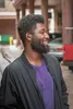A man wearing earbuds and a purple t-shirt, with a black jacket, smiles while looking to the side. Anteprima dell’immagine prodotto