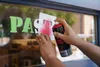 A person is spray painting a letter 'T' onto a window using a stencil and a can of spray paint. Produktbild-Vorschau