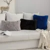 Three fluffy decorative pillows (gray, black, and blue) are arranged on a white sofa, with a white throw blanket draped over the side. A book is open on the sofa in front of the gray pillow. A painting of a deer is visible on the wall behind the sofa. Produktbild-Vorschau
