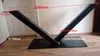 A black V-shaped table base is shown on a wooden floor against a white and light gray wall. Produktbild-Vorschau