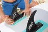 A person's hands are attaching fins to a stand-up paddleboard on a sandy beach. Produktbild-Vorschau