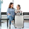 A mother and daughter are sitting on chairs in what appears to be an airport or train station waiting area, with a suitcase between them. Anteprima dell’immagine prodotto