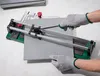 Hands wearing gloves are using a tile cutter on a gray tile. Produktbild-Vorschau