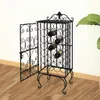 A black metal wine rack with an open door, holding several wine bottles, is placed on a wooden floor against a white wall. A large green leaf plant is visible to the right. Náhľad obrázku produktu