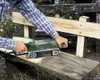 A person is using a Bosch belt sander to sand a wooden bench outdoors. Podgląd zdjęcia produktu