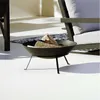 A black metal fire bowl with logs inside, placed on a white patio next to a rug and outdoor furniture. Produktbild-Vorschau