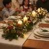 A festive Christmas table setting with a centerpiece of pine branches, candles, and fairy lights. Náhľad obrázku produktu