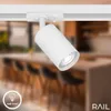 A white track lighting fixture is shown on a white track, with a blurred background of a kitchen or dining area. Produktbild-Vorschau