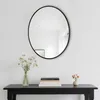 A round mirror with a black frame hangs on a white wall above a black console table. The table holds a vase of flowers, a stack of books, and a small vase with flowers. Produktbild-Vorschau