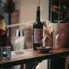 A small, modern LED table lamp is displayed on a wooden shelf next to a bottle of wine and wine glasses, with a blurred background of a person and plants. Anteprima dell’immagine prodotto