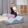A young girl sits on soft play blocks, holding a foam cylinder. The blocks are in various pastel colors and arranged on a carpeted floor. In the background, there are curtains and shelving units. Produktbild-Vorschau