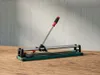 A tile cutter is placed on a wooden surface in front of a concrete wall. Produktbild-Vorschau