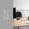 A white LED desk lamp is shown on a wooden desk next to a computer monitor and notebooks. Produktbild-Vorschau