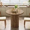 A round wooden dining table with a fluted pedestal base is set in a bright, modern kitchen with a large window overlooking a landscape. Two chairs with beige upholstered seats are positioned at the table, and a woven tray with decorative items sits on the  Produktbild-Vorschau