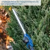 A close-up of a hedge trimmer with a blue handle, cutting a green hedge. Náhľad obrázku produktu
