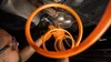 A person wearing orange gloves is working on the underside of a vehicle, using a tool to cut a pipe. Produktbild-Vorschau