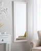 A full-length mirror with a decorative frame hangs on a light gray wall next to a white side table and a chandelier. Náhled obrázku produktu