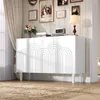 A white sideboard with decorative arched doors is placed in a well-lit room, adorned with artwork and a lamp on top. Voir les images du produit