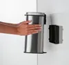 A person's hands are shown reaching for a stainless steel trash can mounted on a white wall. Náhľad obrázku produktu