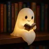 A glowing ghost lamp sits on a wooden shelf in front of a bookshelf. Produktbild-Vorschau