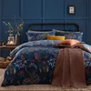 A cozy bedroom scene featuring a bed with a forest-themed duvet cover, pillows, and a throw blanket. The room has dark blue paneled walls and a small wooden table with a lantern and logs. Voir les images du produit