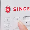 A finger presses a button on a white sewing machine with the 'SINGER' logo visible. Produktbild-Vorschau