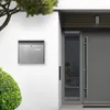A modern mailbox mounted on a white wall next to a grey front door and a green bush. Podgląd zdjęcia produktu