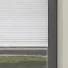 A close-up view of white pleated blinds partially covering a window, with a glimpse of a grassy area outside. Produktbild-Vorschau