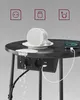 A round black table with integrated power outlets and USB ports, featuring a white cup and saucer on top, and a smartphone charging. Produktbild-Vorschau