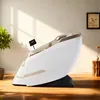 A white and beige massage chair is shown in a well-lit room with wooden floors. Sunlight streams in from a window, casting shadows on the wall. Produktbild-Vorschau