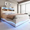 A modern bedroom with a white upholstered bed frame featuring a headboard with built-in shelves and LED lighting. The bed is made up with beige bedding and a throw blanket. The base of the bed also has blue LED lighting. Náhľad obrázku produktu