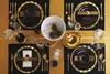 A formal table setting for a meal, featuring black plates with gold trim, gold cutlery, and wine glasses, arranged on a black and gold checkered tablecloth. Anteprima dell’immagine prodotto