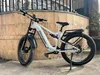 A white electric bicycle is parked on a sidewalk in front of a brick wall and a metal fence. Produktbild-Vorschau