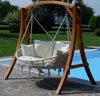 A wooden swing set with a hammock-like seat and cushions, placed on a stone patio next to a swimming pool. Produktbild-Vorschau