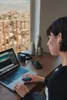 A woman is working on her laptop at a desk by a window, wearing earbuds. Anteprima dell’immagine prodotto