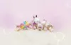 A collection of plush unicorn toys and candy-themed decorations arranged on a fluffy white surface against a soft pink background with whimsical line drawings of candies and clouds. Produktbild-Vorschau