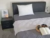 A neatly made bed with a pillow and a white quilted throw. A bedside table with a framed picture and a lit candle is next to the bed. A book and a small bowl of snacks are on the bed. Produktbild-Vorschau