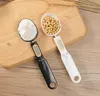Two digital measuring spoons, one black and one white, are placed on a wooden cutting board. The black spoon contains white powder, and the white spoon contains small yellow beans. Both spoons have digital displays. Náhled obrázku produktu