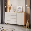 A modern living room scene featuring a beige dresser with gold accents and legs, decorated with a bust, a vase of dried flowers, and a framed artwork. The dresser is placed on a marble floor with curtains in the background. Produktbild-Vorschau