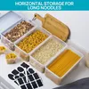 Several clear plastic food storage containers filled with different dry goods like cereal, pasta, oats, and spaghetti are arranged on a white countertop. A wooden cutting board is visible in the background. A sheet of black chalkboard-style labels and a wh Produktbild-Vorschau