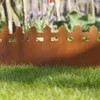 A rusty metal garden border with a picket fence design is placed in a garden, separating green grass from a blurred background of plants and a wooden fence. Produktbild-Vorschau