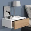 A modern white and wood floating nightstand with an open drawer, mounted on a blue wall next to a bed. A framed picture and a lamp are on top. Produktbild-Vorschau