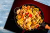 A close-up overhead view of a pan filled with stir-fried vegetables, placed on a surface with a blue wooden texture on the left and a black and red gradient on the right. Náhled obrázku produktu