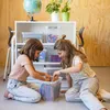Two young girls are sitting on the floor in front of a white shelf, sorting through items in a clear plastic storage box. Produktbild-Vorschau