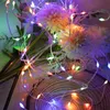 A close-up view of colorful LED string lights intertwined with artificial flowers and greenery, resting on a wooden surface. Produktbild-Vorschau