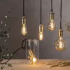 Several Edison-style LED light bulbs with spiral filaments are displayed, some hanging and one inside a glass cylinder, on a wooden surface against a textured gray background. Podgląd zdjęcia produktu