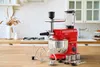 A red kitchen mixer with various attachments and a blender jug, displayed on a wooden countertop in a kitchen setting. Produktbild-Vorschau